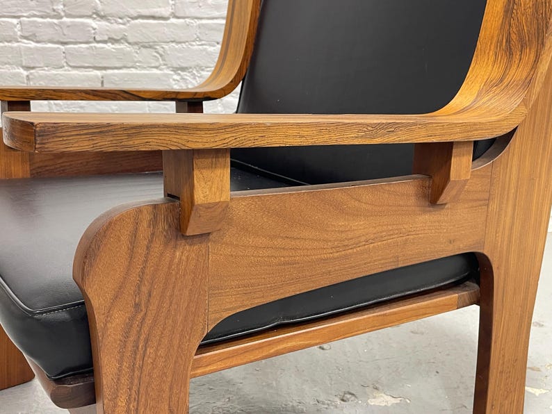 May include: Close-up of a mid-century modern armchair. The chair features a wooden frame with a natural finish and a black leather seat and back. The armrests are curved and the chair has a sturdy, geometric design.