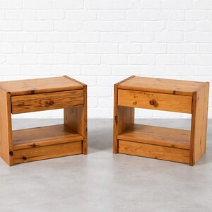 May include: Two matching wooden nightstands. Each nightstand has a drawer with a round wooden knob and an open shelf below. The nightstands are made of light-colored wood and have a simple, rectangular design.
