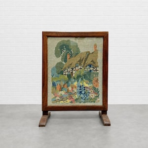 May include: A rectangular fireplace screen featuring a wooden frame and a needlepoint tapestry insert. The tapestry depicts a cottage with a thatched roof surrounded by green trees and a colorful garden filled with various flowers in bloom.