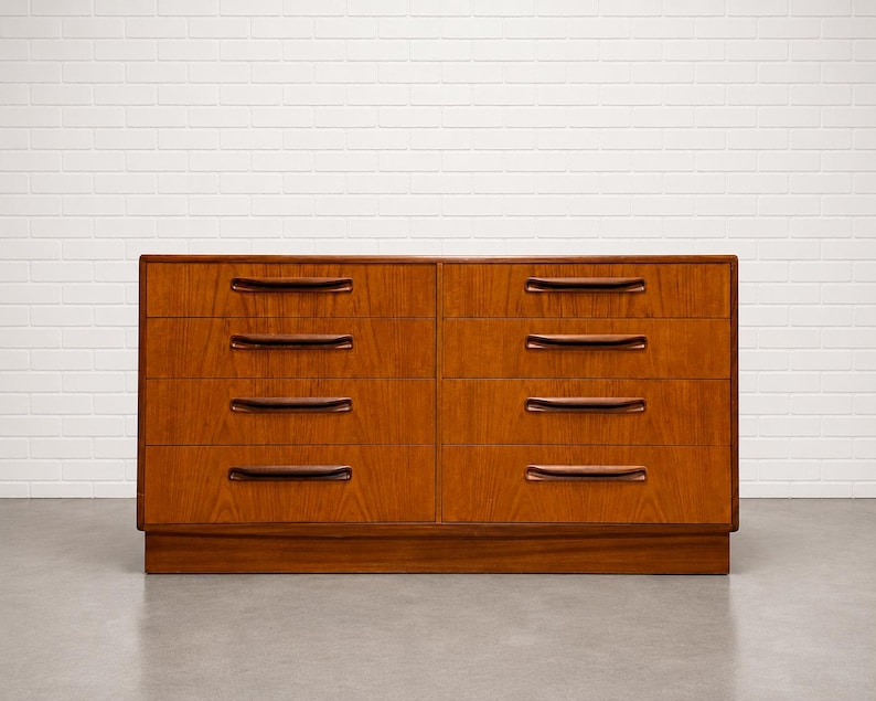 May include: A mid-century modern dresser with six drawers. The dresser is made of wood with a warm, reddish-brown finish. Each drawer has a dark brown, curved handle. The dresser is set against a white brick wall.