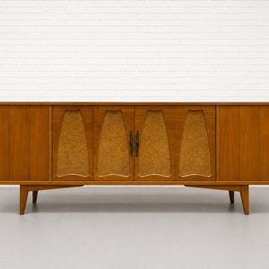 May include: A mid-century modern wooden cabinet with four doors and a low profile. The cabinet is made of a warm-toned wood, with a lighter wood grain pattern on the center doors. It has angled legs and dark metal hardware.