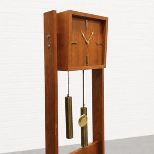 May include: A tall wooden floor clock featuring a square face with gold-toned hands and markers. The clock is suspended within a vertical frame and includes two cylindrical brass weights hanging from chains and a circular brass pendulum.
