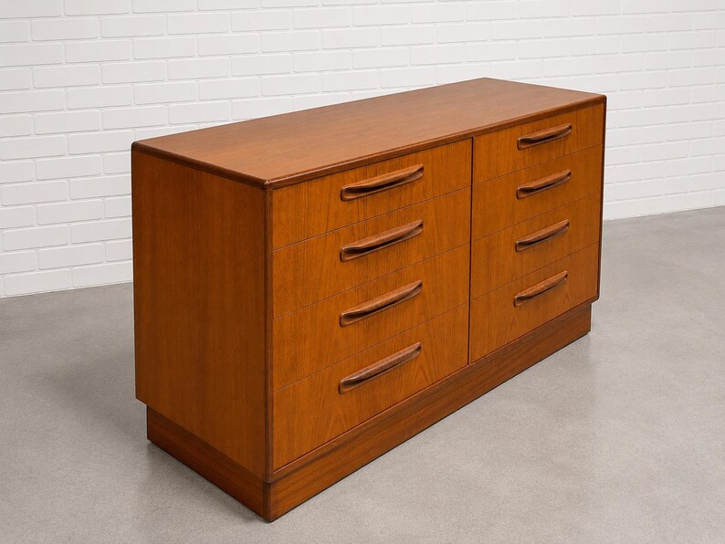 May include: A mid-century modern wooden dresser with eight drawers. The dresser is made of light brown wood and has a rectangular shape. The drawers have curved, recessed handles.