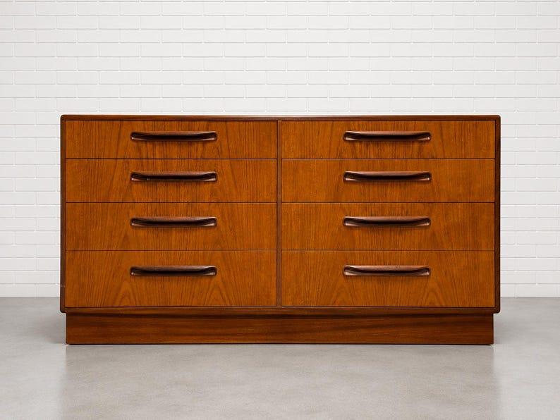 May include: A mid-century modern dresser with six drawers. The dresser is made of wood with a warm brown finish. Each drawer has a dark brown, curved handle. The dresser sits on a low, rectangular base.