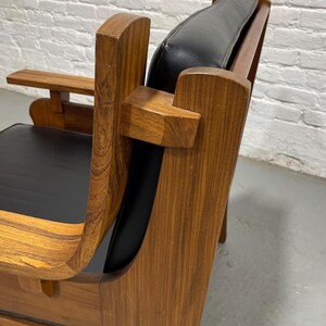 May include: A mid-century modern armchair with a wooden frame and black leather seat and back. The chair features curved armrests and a small side table. The wood has a visible grain.