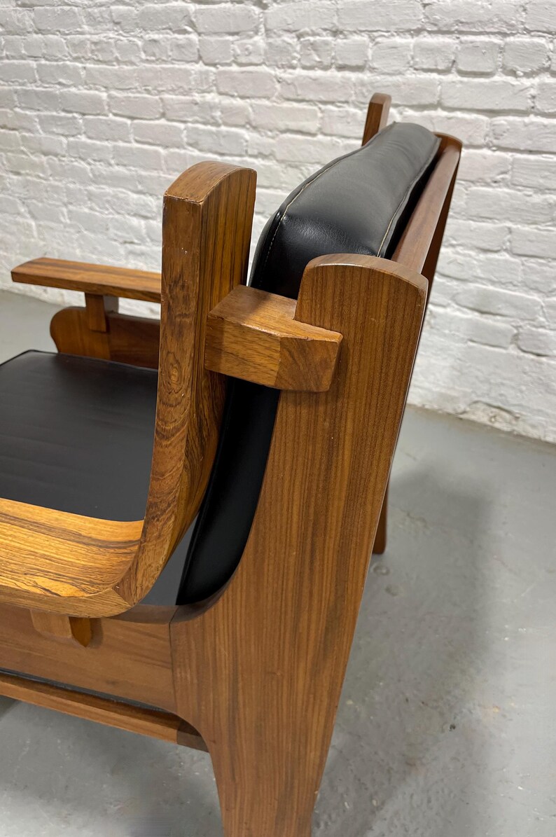May include: A mid-century modern armchair with a wooden frame and black leather seat and back. The chair features curved armrests and a small side table. The wood has a visible grain.