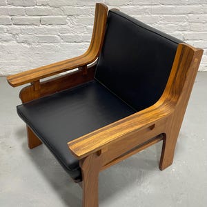 May include: A mid-century modern armchair with a black leather seat and back. The chair features a solid wood frame with curved armrests and legs. The wood has a warm, natural finish, and the chair is set against a white brick wall.