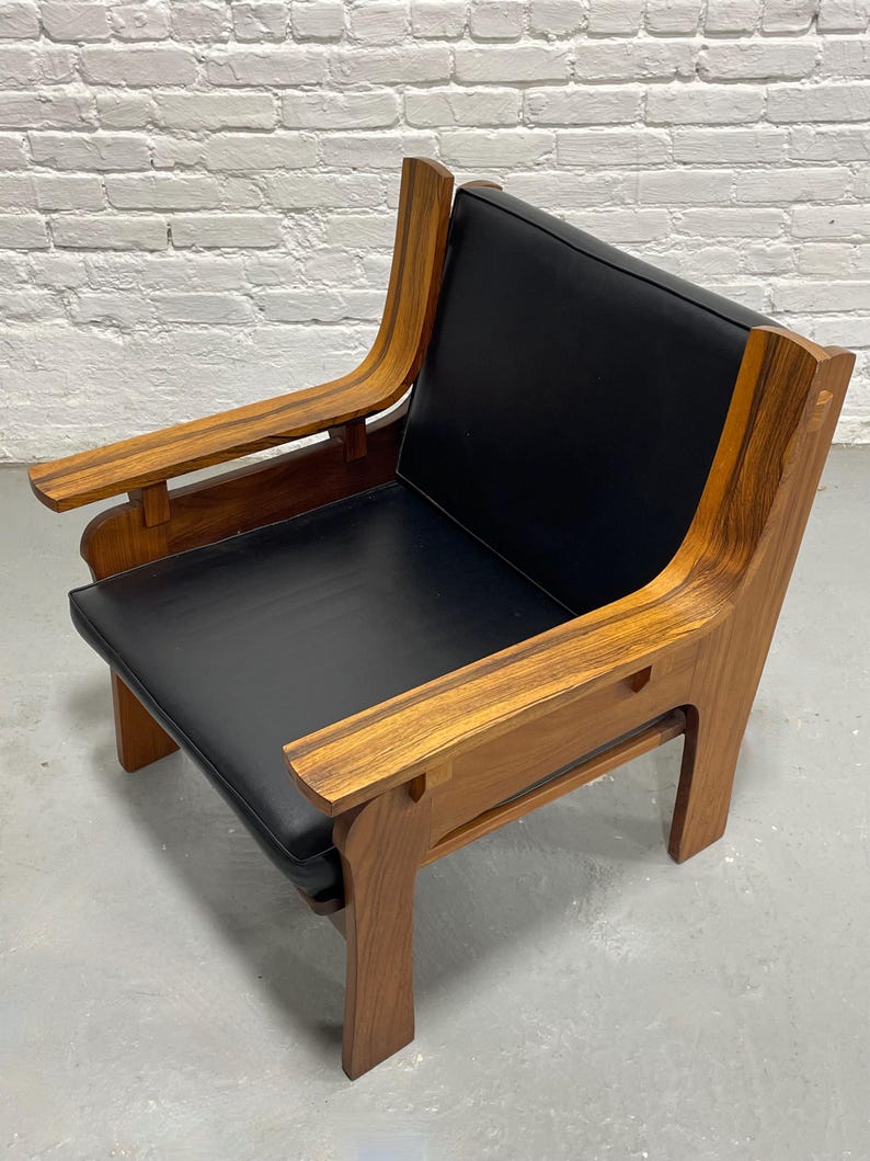 May include: A mid-century modern armchair with a black leather seat and back. The chair features a solid wood frame with curved armrests and legs. The wood has a warm, natural finish, and the chair is set against a white brick wall.
