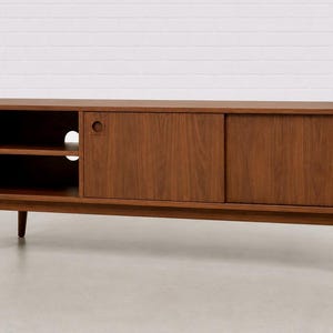 May include: A long, rectangular brown wooden media console featuring four tapered legs, an open shelving unit on the left side with one horizontal shelf, and two sliding cabinet doors on the right side, each with a circular recessed handle.