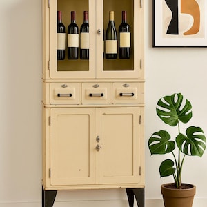 May include: A vintage cream-colored cabinet with glass-fronted upper doors displaying wine bottles. The cabinet has drawers and lower doors, set on black legs with wheels. A potted Monstera plant and a framed abstract artwork complete the scene.