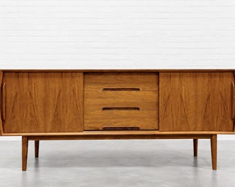 May include: A rectangular wooden sideboard featuring two sliding doors on the left and right sides, each with a vertical handle. The center section contains three stacked drawers with horizontal recessed pulls. The unit stands on four tapered legs.