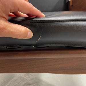 May include: Close-up of a black leather cushion with visible stitching and a small scratch. The cushion is part of a wooden chair, with a dark brown wooden frame visible below.