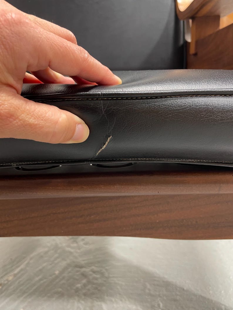 May include: Close-up of a black leather cushion with visible stitching and a small scratch. The cushion is part of a wooden chair, with a dark brown wooden frame visible below.