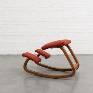 May include: An ergonomic kneeling chair featuring a curved wooden frame and three separate, padded cushions upholstered in a solid rust-orange fabric. The design includes a large seat cushion and two smaller knee pads supported by a continuous base.