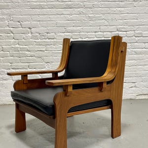 May include: A mid-century modern armchair with a wooden frame and black leather seat and back. The chair has curved armrests and a sturdy, geometric design. The wood has a warm, natural finish, and the chair is set against a white brick wall.