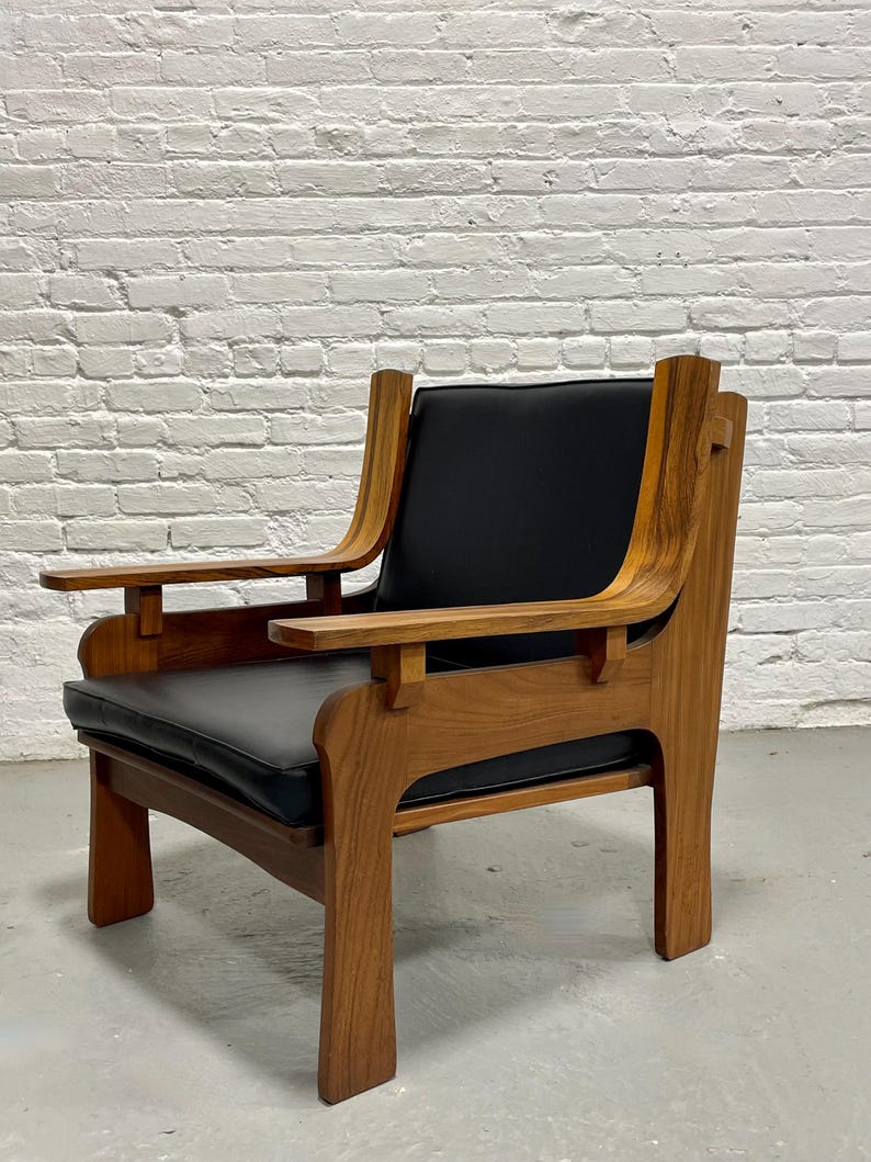 May include: A mid-century modern armchair with a wooden frame and black leather seat and back. The chair has curved armrests and a sturdy, geometric design. The wood has a warm, natural finish, and the chair is set against a white brick wall.