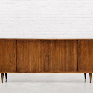 May include: A mid-century modern wooden sideboard with a warm brown finish. The cabinet has two doors on each end and a center section with a ribbed sliding door. It stands on four tapered legs.