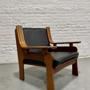 May include: A mid-century modern armchair with a dark wood frame and black leather seat and back. The chair has curved armrests and a rectangular seat cushion. The chair is set against a white brick wall.