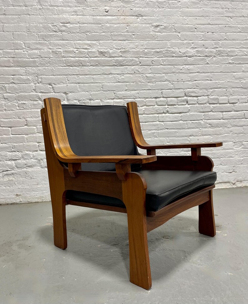 May include: A mid-century modern armchair with a dark wood frame and black leather seat and back. The chair has curved armrests and a rectangular seat cushion. The chair is set against a white brick wall.