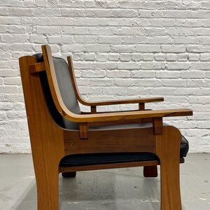 May include: A mid-century modern armchair with a wooden frame and black leather seat and back. The chair has a curved backrest and armrests, and a sturdy, geometric base. The wood has a warm, natural finish.
