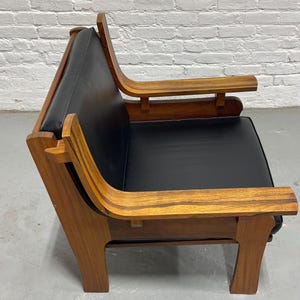 May include: A mid-century modern armchair with a black leather seat and back. The chair features a wooden frame with curved armrests and legs. The wood has a warm, natural finish, and the chair is set against a white brick wall.