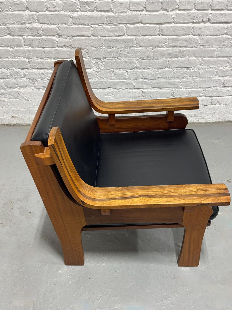 May include: A mid-century modern armchair with a black leather seat and back. The chair features a wooden frame with curved armrests and legs. The wood has a warm, natural finish, and the chair is set against a white brick wall.