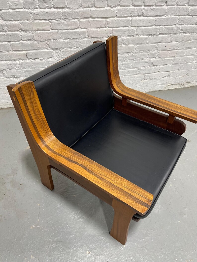 May include: A mid-century modern armchair with a black seat and backrest. The chair features curved wooden armrests and a wooden frame with a warm, natural finish. The design is minimalist and stylish, suitable for a living room or lounge area.