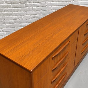 May include: A mid-century modern dresser with a warm, reddish-brown wood finish. The dresser has six drawers with recessed pulls. The top surface is smooth and polished, reflecting the light. The piece is a classic example of vintage furniture.