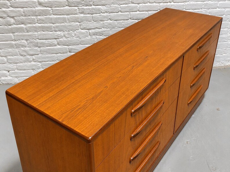 May include: A mid-century modern dresser with a warm, reddish-brown wood finish. The dresser has six drawers with recessed pulls. The top surface is smooth and polished, reflecting the light. The piece is a classic example of vintage furniture.