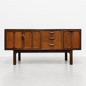 May include: A long wooden sideboard featuring a rich brown finish, two cabinet doors with recessed panels on the left, three central drawers with horizontal handles, and one cabinet door on the right, all supported by four sturdy wooden legs.