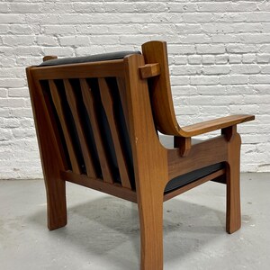 May include: A mid-century modern armchair with a wooden frame and black leather seat and back. The chair has a slatted back design and angled armrests. The wood has a warm, natural finish.