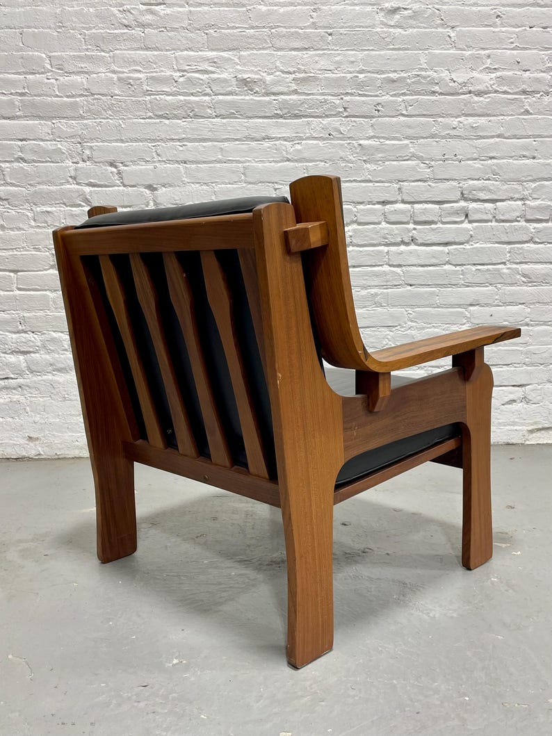 May include: A mid-century modern armchair with a wooden frame and black leather seat and back. The chair has a slatted back design and angled armrests. The wood has a warm, natural finish.