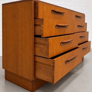 May include: A mid-century modern dresser with eight drawers, crafted from warm-toned wood. The drawers feature integrated, curved handles. Several drawers are open, revealing the dovetail joinery. The dresser sits on a low, rectangular base.