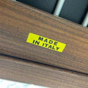 May include: Close-up of a wooden furniture component with a yellow sticker that reads "MADE IN ITALY" in black text. The wood has a rich, dark brown color and visible grain.
