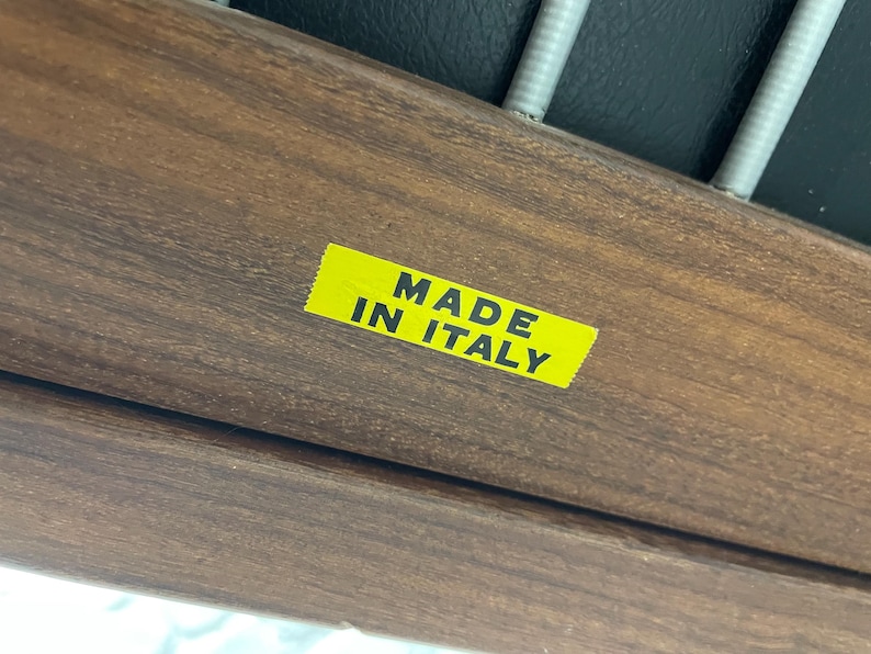May include: Close-up of a wooden furniture component with a yellow sticker that reads "MADE IN ITALY" in black text. The wood has a rich, dark brown color and visible grain.
