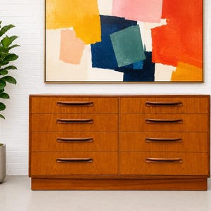 May include: A mid-century modern wooden dresser with eight drawers and dark brown handles. The dresser is made of light brown wood and sits on a low base. Above the dresser is an abstract painting with yellow, pink, blue, and red colors.