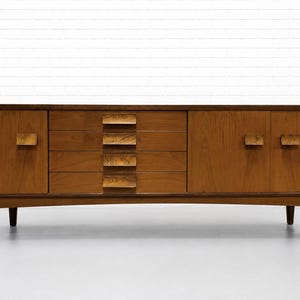 May include: A long wooden sideboard featuring a rich brown wood grain finish. The piece includes two side cabinets with rectangular handles and a central column of four horizontal drawers, each equipped with a matching wooden pull. It stands on four legs.
