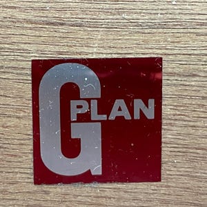 May include: A rectangular red sticker with the word "PLAN" in white text and a large silver "G" on the left side. The sticker is on a light brown wooden surface.