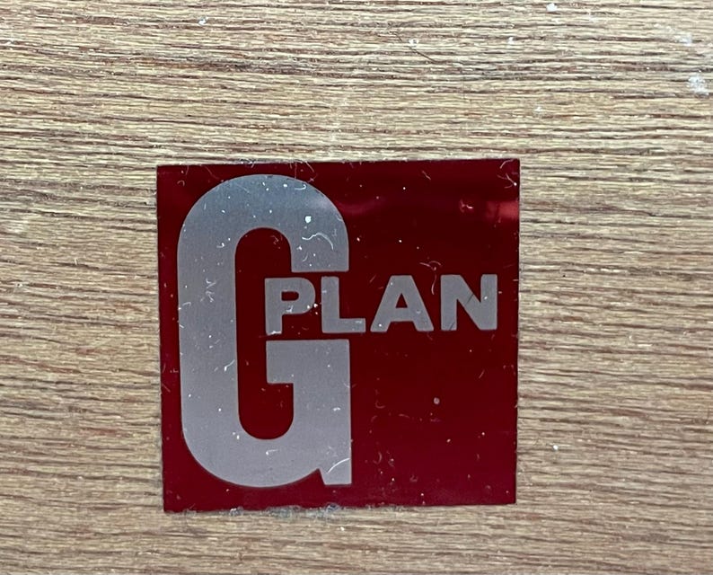 May include: A rectangular red sticker with the word "PLAN" in white text and a large silver "G" on the left side. The sticker is on a light brown wooden surface.