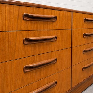 May include: A mid-century modern dresser with six drawers. The dresser is made of wood with a warm, reddish-brown finish. Each drawer has a curved, recessed handle. The dresser is set against a white brick wall.