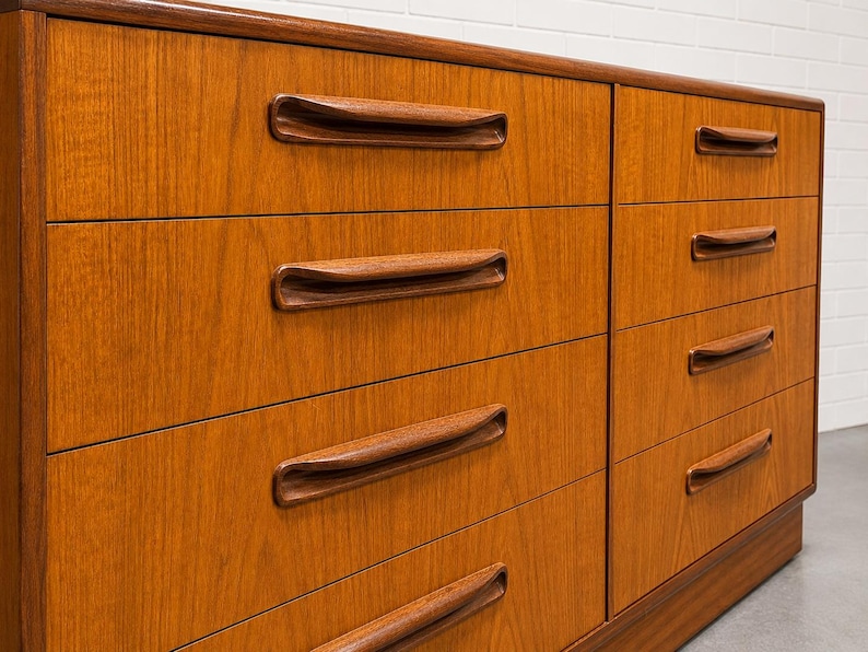 May include: A mid-century modern dresser with six drawers. The dresser is made of wood with a warm, reddish-brown finish. Each drawer has a curved, recessed handle. The dresser is set against a white brick wall.
