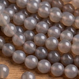May include: A close-up of grey gemstone beads, round and polished. The beads range in shade from light to dark grey, with a translucent appearance. Some beads are loose, others are strung together, on a wooden surface.