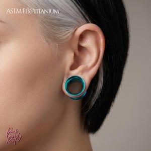 Può includere: Un tunnel per orecchio circolare color verde acqua realizzato in titanio ASTM F136. Il gioiello presenta un centro cavo liscio e una finitura metallica lucida, inserito nel lobo dell'orecchio. Il testo ASTM F136 TITANIUM è visibile in alto a sinistra.