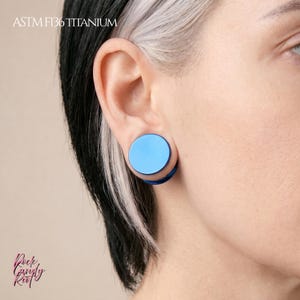 May include: A bright blue circular ear plug made of ASTM F136 titanium. The jewelry features a smooth, flat, solid blue front surface and is inserted into an earlobe. The text ASTM F136 TITANIUM is visible in the upper left corner of the image.