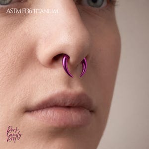 Pode incluir: Um anel de septo fúcsia metálico com um design de presa curva e cônica usado no nariz. O texto ASTM F136 TITANIUM é visível no topo e o texto Rock Candy Riot é visível no canto inferior esquerdo da imagem.