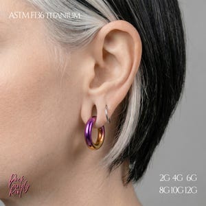 May include: A smooth, circular hoop earring made of ASTM F136 titanium with a gradient finish transitioning from purple to gold. The jewelry is worn in an earlobe piercing, accompanied by a small silver ring. Text reads ASTM F136 TITANIUM and 2G 4G 6G 8G 10G 12G.