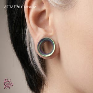 Puede incluir: Un túnel de oreja de titanio ASTM F136 iridiscente con forma circular y un acabado liso y pulido que cambia entre tonos verdes, morados y azules. La joya está insertada en el lóbulo de la oreja y muestra un centro hueco.