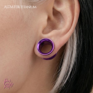 Peut inclure: Un tunnel d'oreille en titane violet au design circulaire et lisse. Le bijou porte la mention ASTM F136 TITANIUM et présente un centre creux. La pièce est portée dans un lobe d'oreille étiré, mettant en valeur une finition métallique vive.