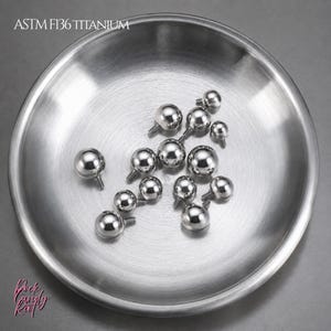 May include: A collection of small, polished, spherical ASTM F136 titanium body jewelry balls with threaded posts, resting inside a circular, brushed metal bowl. The text ASTM F136 TITANIUM is visible in the upper left corner of the image.