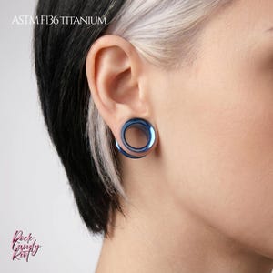 May include: A metallic blue ear tunnel plug with a smooth, circular design inserted into an earlobe. The text ASTM F136 TITANIUM is visible in the upper left corner, and the text Rock Candy Riot is visible in the lower left corner.